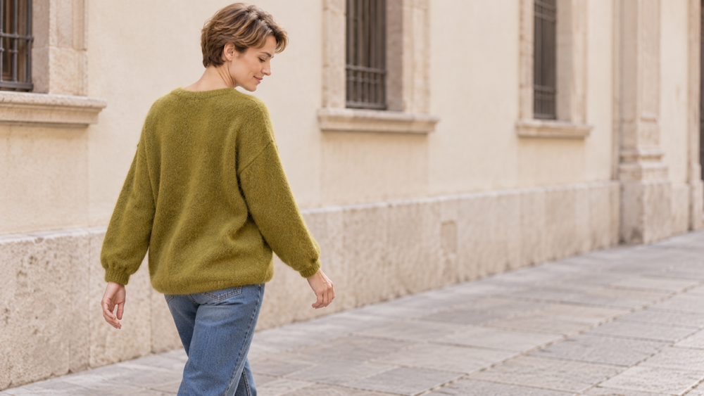 Su un marciapiede cittadino, una donna cammina con un maglione ampio e jeans dritti, ripresa di tre quarti da dietro.